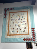 A quilt hanging on a white wall featuring an alphabet panel with three borders. The outermost blue border features red geometric flying geese accents. 