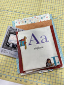 A stack of the fabrics included in the Alphabet Panel kit, including the panel. The pattern for this kit is included alongside them. 