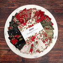 A charm pack from Moda's Candlelight Christmas collection displayed atop a white plate on a wooden table. 