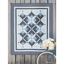 A quilt hanging on a wall featuring various log cabin blocks in blue. The wall features weathered wood paneling, and a blue bike is parked nearby. 
