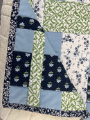 A close-up of a handmade quilted patchwork blanket featuring various fabric prints including green and white geometric patterns, navy blue floral prints, and solid light blue squares.