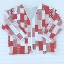 A quilted jacket featuring a patchwork red and white design. It is laid out on a white wood background. 